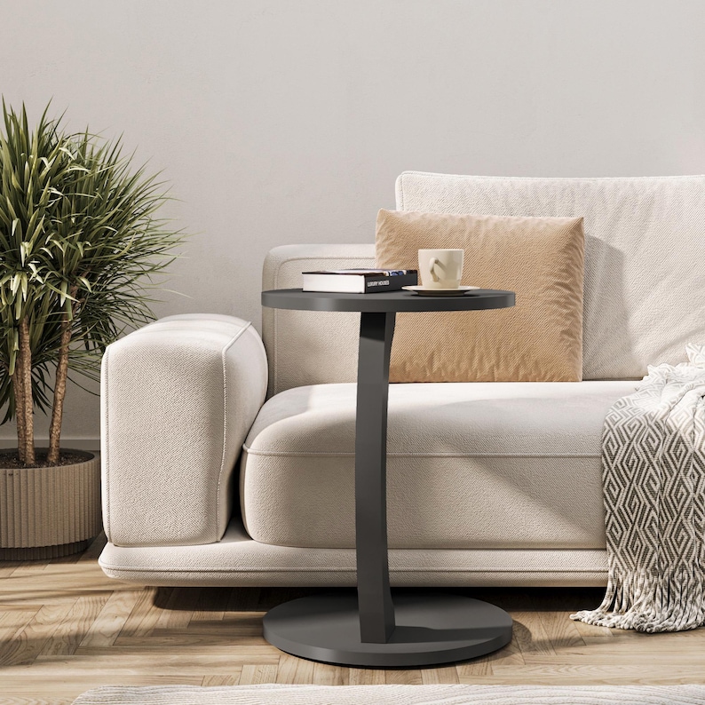 Puede incluir: Una mesa auxiliar de color gris oscuro con una tapa redonda y una pata curva. La mesa est&aacute; al lado de un sof&aacute; blanco con un coj&iacute;n beige y una manta.