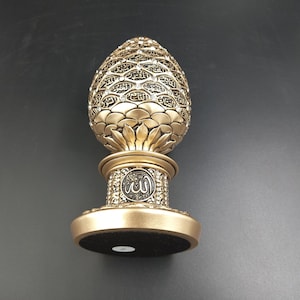 Pode incluir: Um objeto decorativo dourado em forma de ovo com desenhos intrincados e a palavra "Allah" inscrita na base. O ovo &eacute; coberto por pequenos pontos em relevo e tem uma base preta.