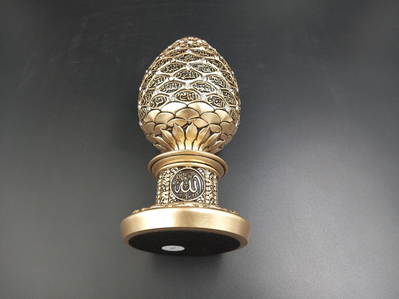 Pode incluir: Um objeto decorativo dourado em forma de ovo com desenhos intrincados e a palavra "Allah" inscrita na base. O ovo &eacute; coberto por pequenos pontos em relevo e tem uma base preta.