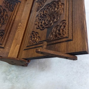 May include: A wooden book stand with intricate floral carvings. The stand is made of dark brown wood and features a hinged design for adjustability. The carved details add a decorative element to this functional item, suitable for holding books or displaying items.