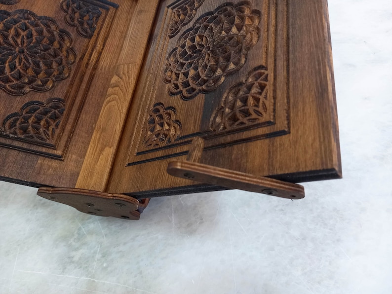 May include: A wooden book stand with intricate floral carvings. The stand is made of dark brown wood and features a hinged design for adjustability. The carved details add a decorative element to this functional item, suitable for holding books or displaying items.