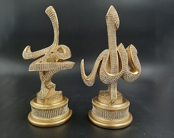 Pode incluir: Duas estatuetas decorativas douradas com as palavras árabes "Allah" e "Muhammad" escritas em strass. As estatuetas estão sobre um fundo preto.