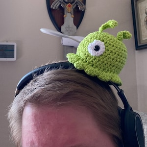 Op de afbeelding: Een groene gehaakte slak met een wit oog en een zwarte pupil zit op een zwarte koptelefoon. De koptelefoon zit op het hoofd van een persoon.