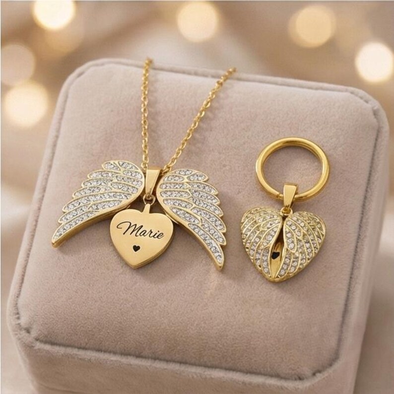 May include: A gold-colored necklace and keychain set. The necklace features a heart pendant with the name "Marie" and wing accents, embellished with clear crystals. The keychain has a heart-shaped pendant with wing details.