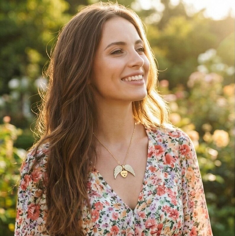 May include: A woman wearing a gold necklace with a heart-shaped pendant and angel wings. The pendant has a small heart in the center. She is wearing a floral print dress with a V-neck. The background is a garden with soft sunlight.