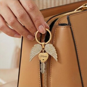 May include: A gold-colored keychain with angel wings and a heart-shaped charm. The heart charm is engraved with the name "Marie". The keychain is attached to a key and a brown leather handbag.