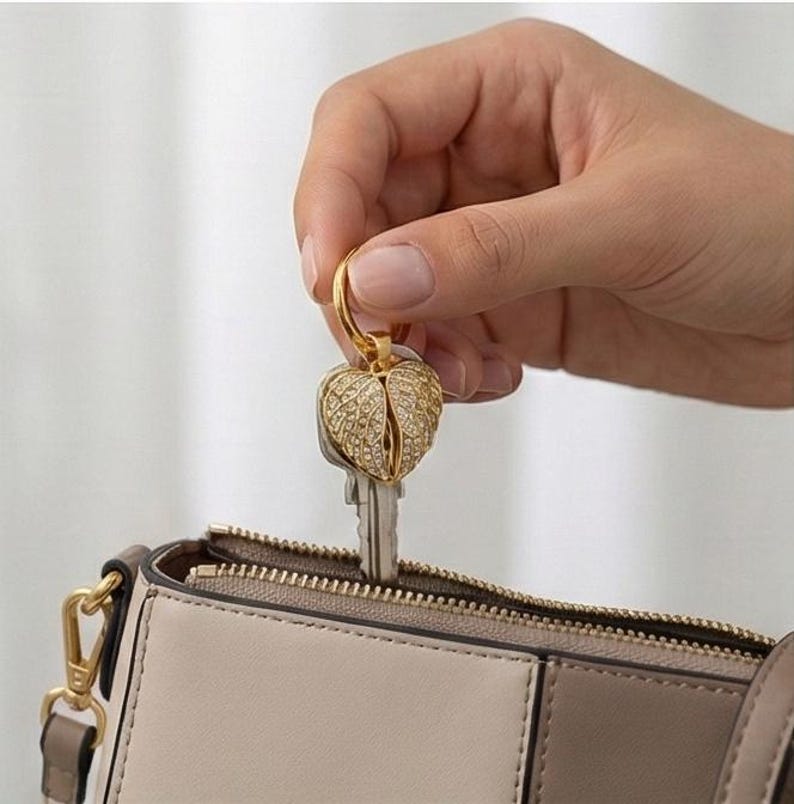 May include: A gold-colored key ring with a heart-shaped charm encrusted with small, clear crystals. The key ring is attached to a silver key. The key and key ring are held near a beige and brown purse.
