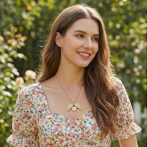 May include: A gold-colored necklace with a heart-shaped pendant and angel wings. The pendant has the word "Mom" engraved on it. The woman is wearing a floral print dress with a square neckline.