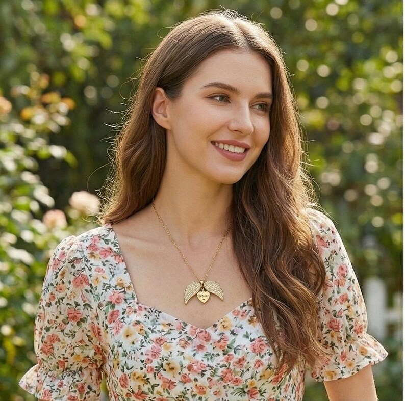 May include: A gold-colored necklace with a heart-shaped pendant and angel wings. The pendant has the word "Mom" engraved on it. The woman is wearing a floral print dress with a square neckline.
