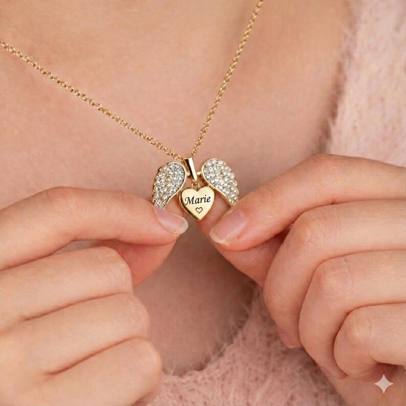 May include: Gold-colored necklace with a heart-shaped pendant and angel wings. The heart pendant is engraved with the name "Marie" and a small heart symbol. The wings are encrusted with small, clear crystals.