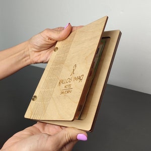 Puede incluir: Una cubierta de menú de madera con un acabado marrón claro. La cubierta tiene una solapa que se abre para revelar un bolsillo para guardar menús u otros materiales. La cubierta está grabada con el texto "KALLOS IMAR HOTEL SANTORINI".