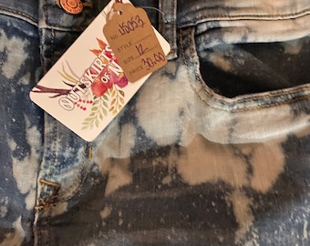 May include: A pair of blue denim jeans with a bleach-splattered design. The jeans have a tag attached that says "Outskirts of Nowhere" and has a price tag of $30.00. The tag also says "No. 16063, Style 81, Size 12."