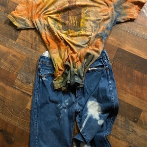 May include: A tie-dye t-shirt with the words "Junk Gypsy" printed on the front. The shirt is paired with blue jeans that have been bleached with white spots.