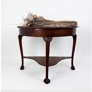 May include: A dark brown, antique-style, round side table with a decorative edge. The table has three carved legs and a lower shelf. A bundle of dried flowers rests on the tabletop. The table is approximately 30 inches in diameter.