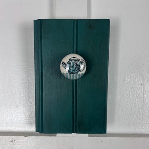 May include: A teal-colored wooden shutter with a white and blue ceramic knob. The knob features a teapot design. The shutter is mounted on a white surface.