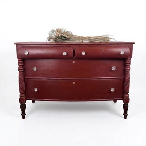 May include: A burgundy-colored antique dresser with three drawers and crystal knobs. The dresser has decorative carved accents on the legs and sides. A bundle of dried flowers rests on top of the dresser.