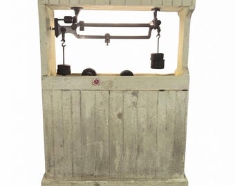 May include: A pale green wooden scale housing with a vertical plank design and a wide top ledge. A dark metal balance scale mechanism is suspended within the open upper frame, featuring two hanging weights and a small circular red logo.