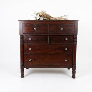 May include: A dark brown wooden dresser with four drawers and two smaller drawers at the top. The dresser has crystal knobs and decorative turned legs. A dried floral arrangement rests on top.