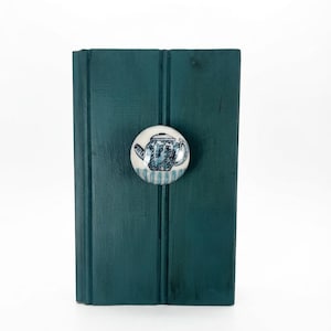 May include: A round white ceramic knob featuring a blue and white illustration of a teapot, mounted on a rectangular dark teal wooden board with vertical grooves. The knob has a smooth, glossy finish and a simple, functional design.
