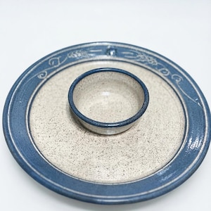 May include: A round ceramic serving dish with a speckled cream-colored center and a wide blue rim. A small, matching bowl is attached to the center of the plate. The blue rim features a subtle, etched wheat pattern around the entire circumference.