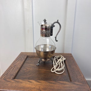 May include: A silver and glass electric coffee pot with a cord on a wooden surface.