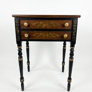 May include: A small, two-drawer side table with a black base and legs. The drawers are wood-toned with gold floral accents and gold knobs. The table's top is dark brown, and the legs have gold detailing.