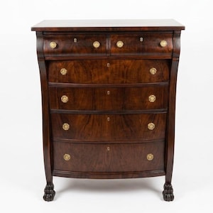 May include: A dark brown wooden dresser with six drawers and gold-colored knobs. The dresser has a curved front and rests on claw feet. The wood grain is visible, and the top is a lighter shade of brown.