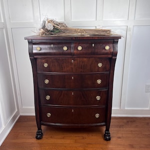 May include: A dark wood dresser with five drawers and gold-colored knobs. The dresser has a curved front and rests on claw feet. A decorative arrangement of dried flowers and grasses sits on top.