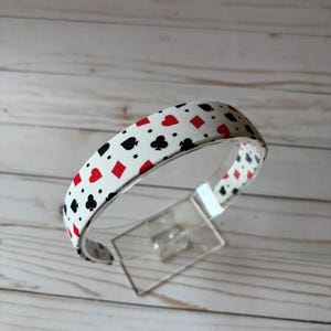 May include: A white headband with a repeating pattern of playing card suits in red and black. The headband is displayed on a clear plastic stand against a light wood background.