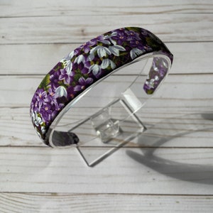 May include: Purple and white floral headband with a green background. The headband is made of fabric and is designed to be worn around the head.