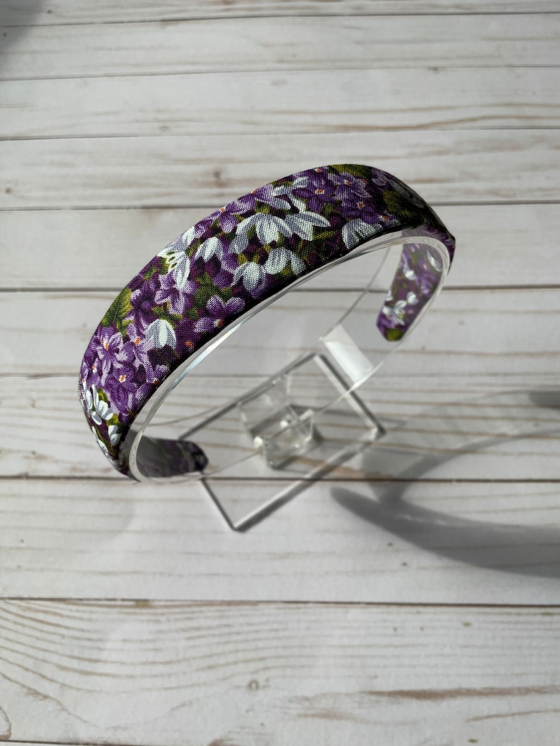May include: Purple and white floral headband with a green background. The headband is made of fabric and is designed to be worn around the head.