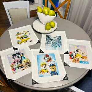 May include: Five personalized white canvas placemats with black trim and names printed on the bottom. The placemats feature images of a construction truck, a yeti, a group of kittens, a group of dogs wearing party hats, and a group of Paw Patrol characters.