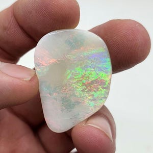 Puede incluir: Una piedra preciosa de ópalo pulida en forma de pera. El ópalo muestra un color base blanco lechoso con destellos iridiscentes de verde, azul y rojo en su superficie. La piedra se sostiene entre los dedos, mostrando su belleza natural y su juego de colores único.