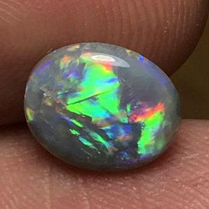 May include: A polished, oval-shaped opal gemstone. The opal displays a dark base color with vibrant flashes of green, blue, and red iridescence. The stone is held between fingers, showcasing its natural beauty and play of color.
