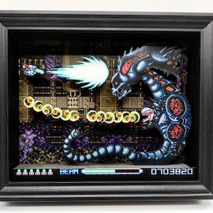 May include: A 3D pixel art diorama of a video game scene featuring a blue and red alien-like creature with a large mouth and a long tail. The creature is firing a blue beam at a small spaceship. The diorama is framed in black and includes a pixelated game screen with the text "BEAM" and a score of "0103820".