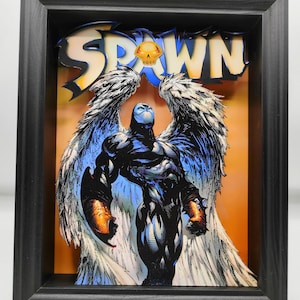 Puede incluir: Una pieza de arte pop-up 3D enmarcada con Spawn, un personaje de cómic, con las alas extendidas. El fondo es un degradado de naranja y amarillo. El título "Spawn" está en letras grandes y en negrita sobre el personaje.