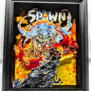 Può includere: Opera d'arte incorniciata con la scritta "SPAWN" sopra un'illustrazione dettagliata di fumetti. L'opera è in una cornice nera e mostra una scena infuocata con colori vivaci. Effetto 3D.