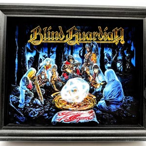 May include: Framed artwork featuring the band Blind Guardian's logo in gold lettering. The image depicts a fantasy scene with figures gathered around a glowing orb. The frame is a dark gray color.