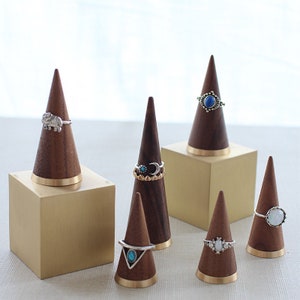 May include: Six wooden ring display cones with gold trim, each with a different ring on top. The cones are arranged on a gold square base and a white surface.
