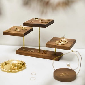 May include: A tiered wooden jewelry stand with gold metal rods. The stand has three square shelves and a round base. The shelves are filled with gold rings, earrings, and bracelets.