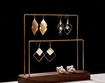 May include: A gold metal earring stand with a wooden base. The stand has two tiers for holding earrings. There are six pairs of earrings on the stand, including gold, black, and white earrings.