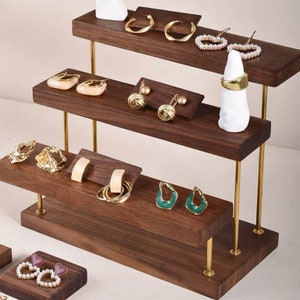 May include: A three-tiered wooden jewelry display stand with gold metal accents. The stand features a variety of earrings, rings, and necklaces.