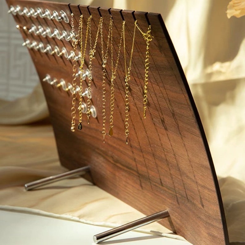 Puede incluir: Un expositor de joyas de madera con un dise&ntilde;o curvo y patas de metal. El expositor tiene m&uacute;ltiples ganchos para colgar collares y una secci&oacute;n con alfileres de pl&aacute;stico transparente para sujetar pendientes.
