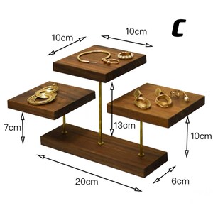 May include: A three-tiered wooden jewelry stand with gold metal rods. The stand is 20 cm wide, 13 cm tall, and 10 cm deep. The top tier is 10 cm wide, the middle tier is 10 cm wide, and the bottom tier is 6 cm wide. The stand is perfect for displaying rings, earrings, necklaces, and bracelets.
