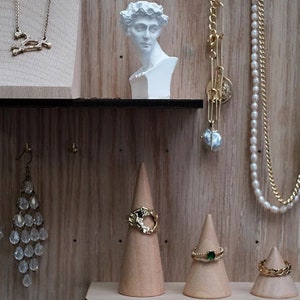 May include: A wooden display with a white plaster bust of a man, a gold chain necklace with a skull pendant, a gold chain necklace with a pearl pendant, a pair of crystal earrings, and three wooden ring cones with gold rings.