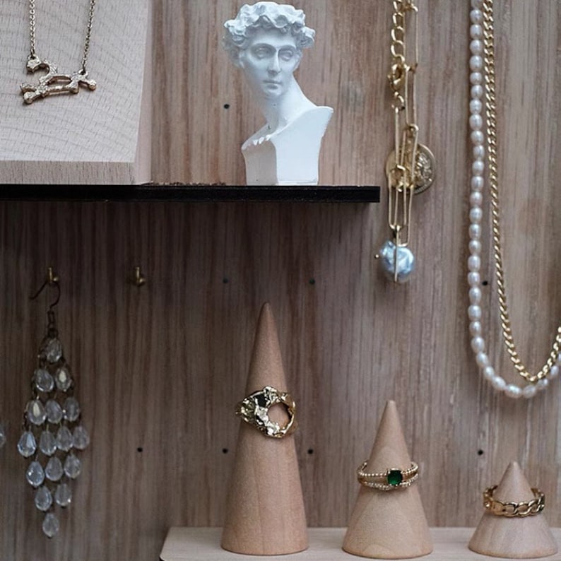 May include: A wooden display with a white plaster bust of a man, a gold chain necklace with a skull pendant, a gold chain necklace with a pearl pendant, a pair of crystal earrings, and three wooden ring cones with gold rings.