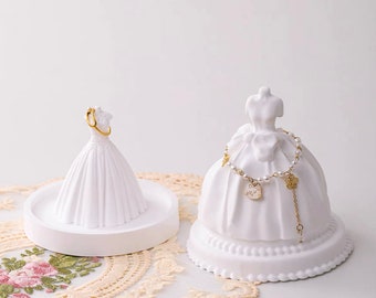 Puede incluir: Dos soportes para anillos y pulseras de cerámica blanca con forma de vestidos de novia. El soporte más grande tiene una pulsera de perlas con abalorios. El soporte más pequeño tiene un anillo de oro.
