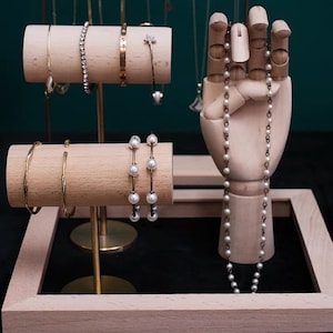 May include: A wooden jewelry display stand with three wooden dowels for bracelets and a wooden hand holding a pearl necklace. The stand is on a black surface inside a wooden frame.