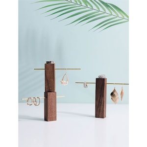 May include: A wooden jewelry stand with two gold metal bars for hanging earrings. The stand is made of dark brown wood and has a modern design.