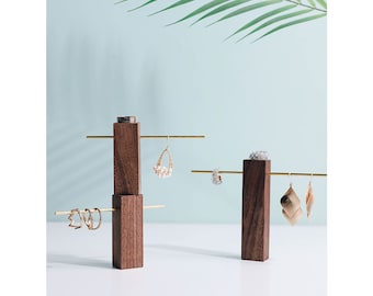 May include: A wooden jewelry stand with two gold metal bars for hanging earrings. The stand is made of dark brown wood and has a modern design.
